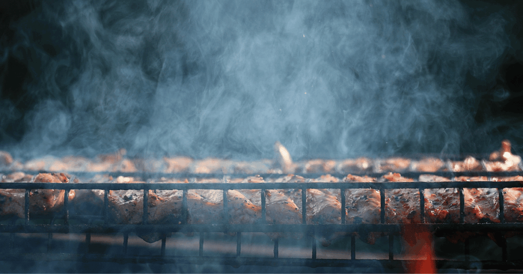 Smoked meats cooking over a low fire with rich wood smoke rising at John Mull’s Meats