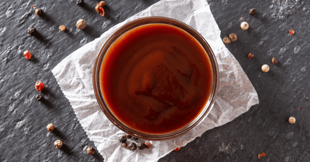 Top-down view of rich BBQ sauce in a glass bowl surrounded by whole peppercorns